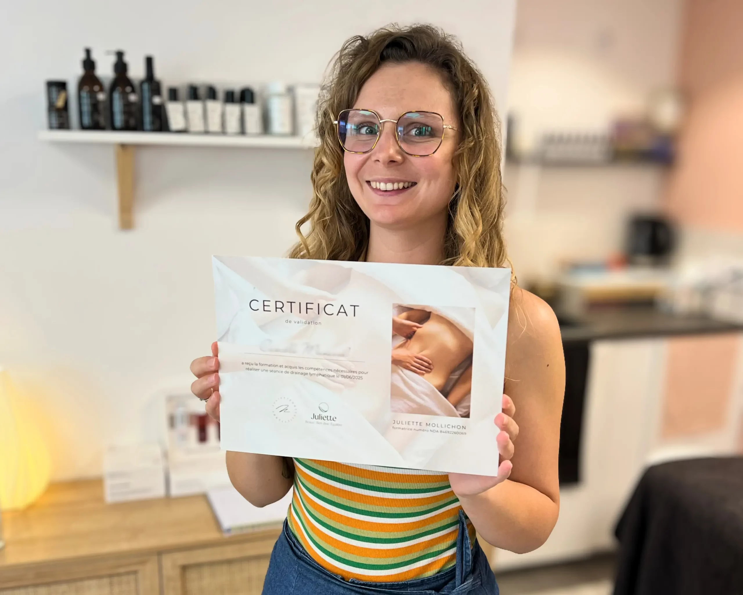 Femme souriante tenant son certificat de validation en drainage lymphatique dans un institut de beauté, avec une étagère de produits cosmétiques en arrière-plan.